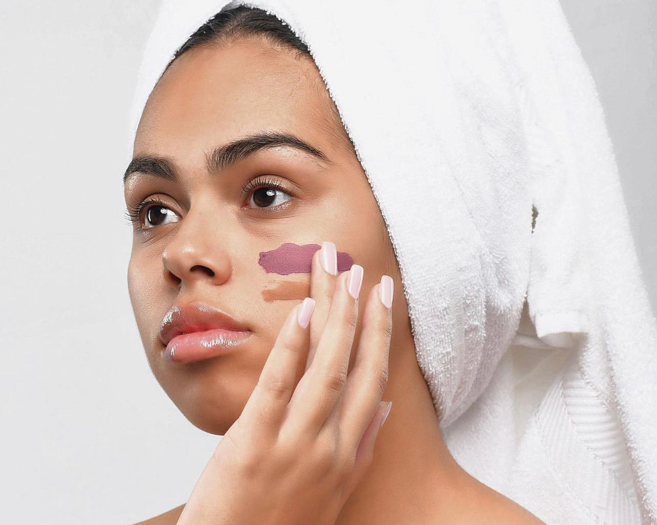 A tan skinned woman wearign a white towel around her hair, wearing a natural facial on her cheek while she touches it with her hand