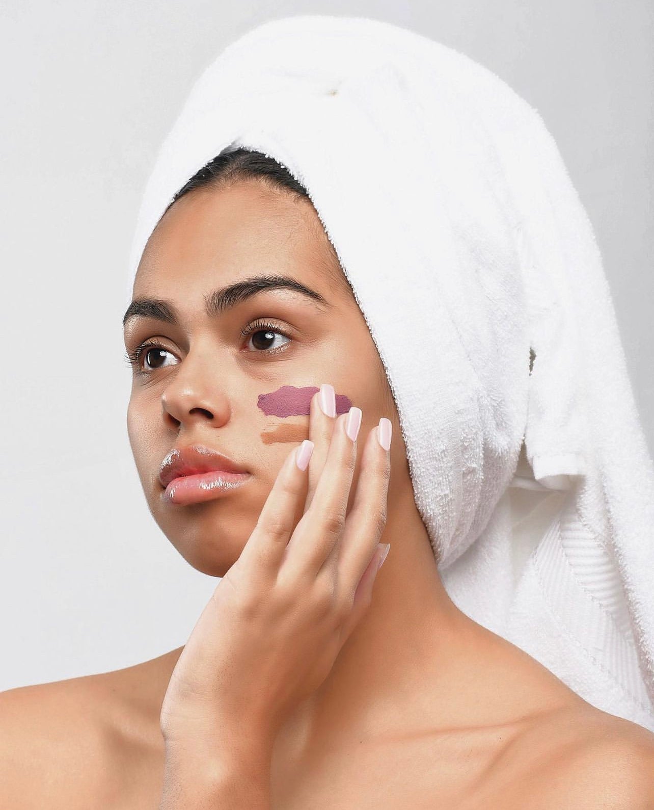 A tan skinned woman wearign a white towel around her hair, wearing a natural facial on her cheek while she touches it with her hand