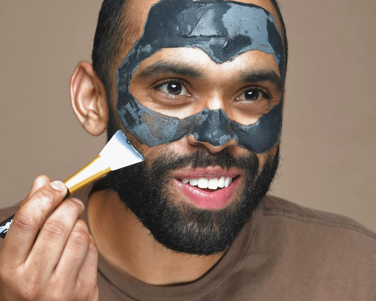 LATINO MAN PUTTING ON CHARCOAL LAVA SALT DRY MASK, A NATURAL FACIAL WITH A BROWN SHIRT